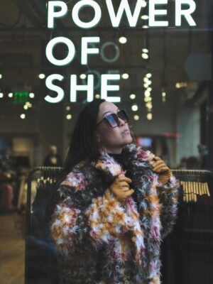 Woman in fur coat poses confidently by 'Power of She' sign indoors.