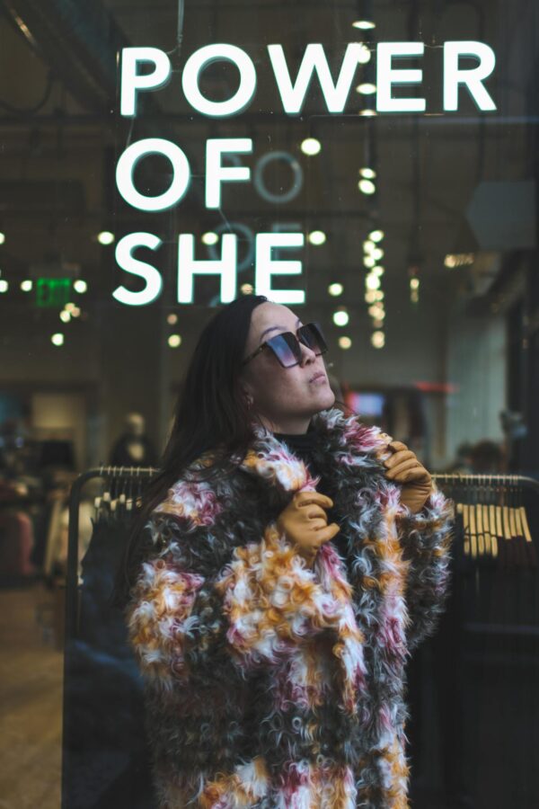 Woman in fur coat poses confidently by 'Power of She' sign indoors.